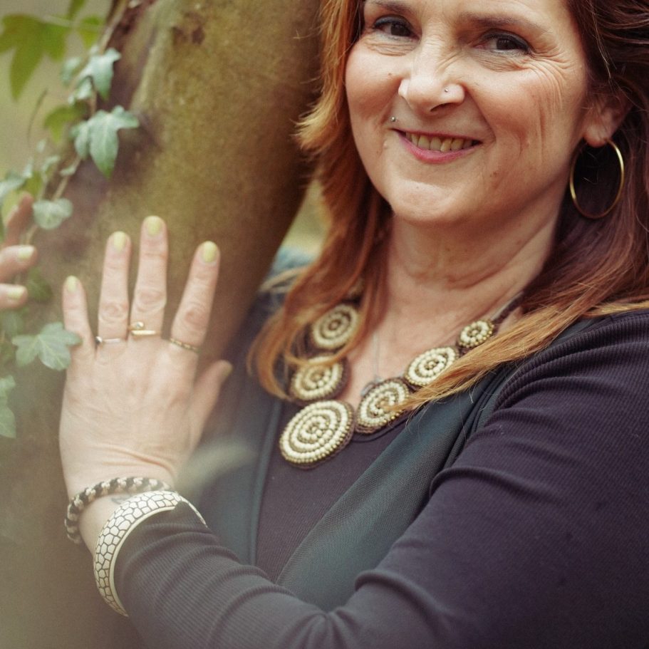 Dominique Bridstrup Frau mit langen roten Haaren, die an einem Baum steht und lächelt.